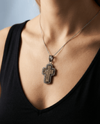 Close-up of authentic 17th–18th century sand-cast copper Old Believers Orthodox cross with historical aged patina, set in oxidized sterling silver with natural diamond accents on a person neck